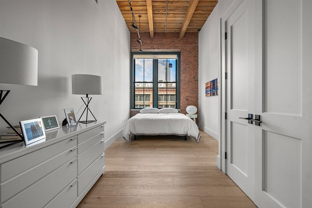 This is a bright and airy primary bedroom featuring hardwood floors, exposed brick, and a large window providing natural light. The room is minimally furnished with a bed, side table, and a dresser with lamps and picture frames. The overall style is modern and loft-like, creating a comfortable and inviting space.