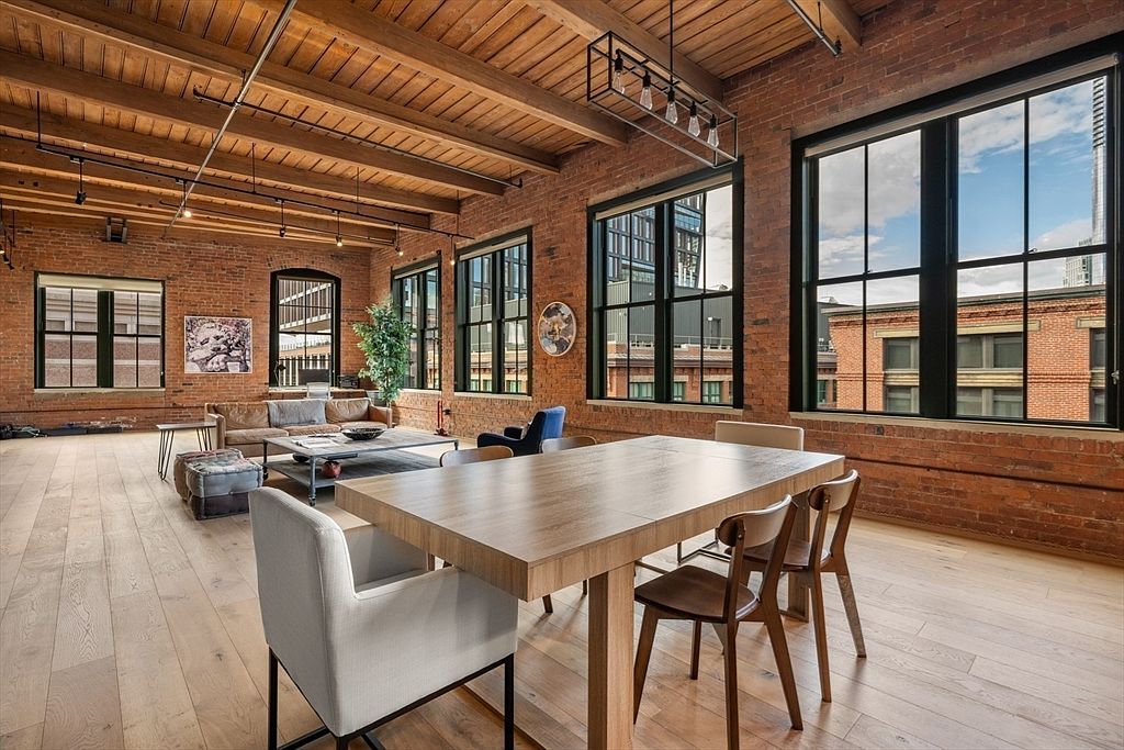 This interior shot showcases a spacious dining area within a loft-style apartment. The room features exposed brick walls, large windows offering city views, and a wooden beam ceiling. A large wooden dining table with modern chairs dominates the foreground, while a living area with a sofa and coffee table is visible in the background, creating an open and inviting atmosphere.