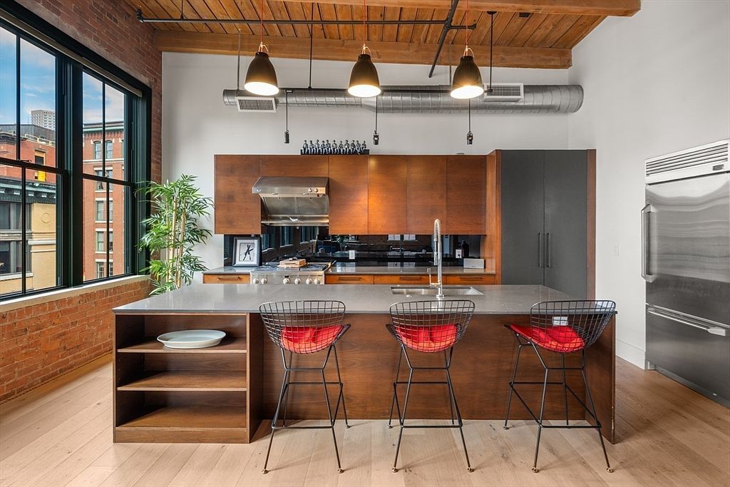 This is a modern kitchen with a large island featuring seating for three. The kitchen has wooden cabinets, stainless steel appliances, and pendant lighting. A large window provides natural light and a view of the city.