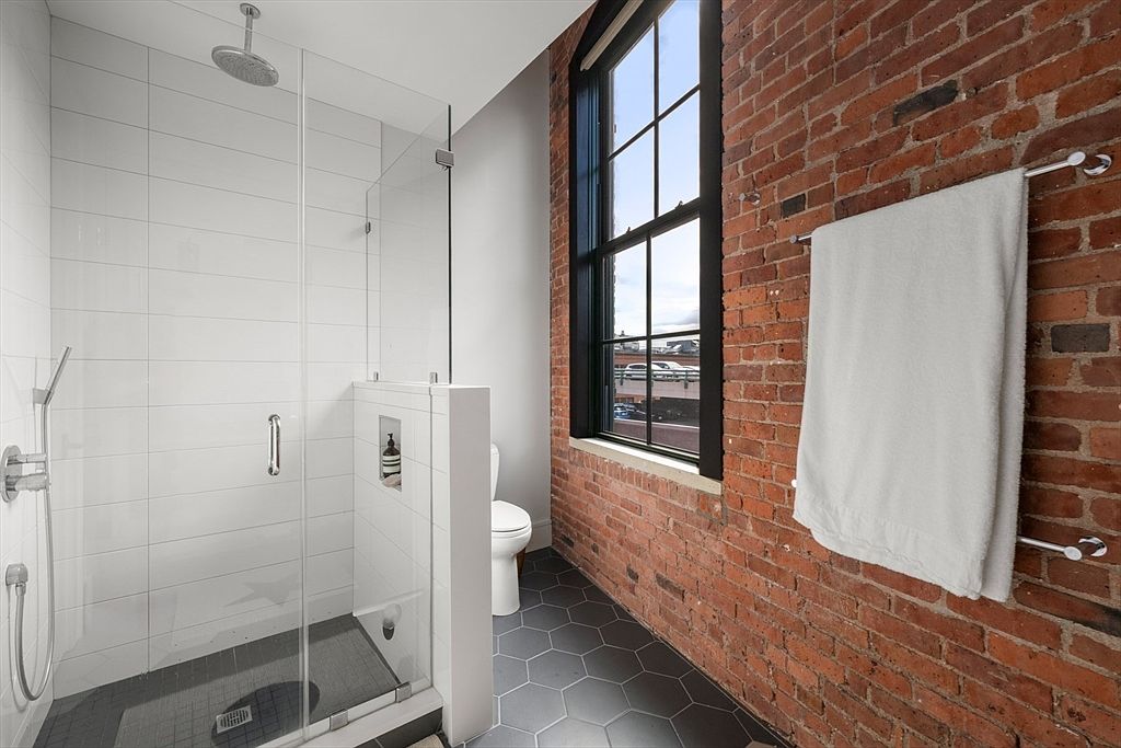 This is a modern primary bathroom featuring a glass-enclosed shower with white subway tile and a rainfall showerhead. The room has a brick accent wall with a towel rack and a large window providing natural light. The flooring is a dark gray hexagonal tile, and a toilet is positioned next to the shower.
