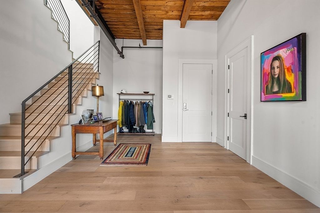 This interior shot showcases a modern hallway with wooden floors and a staircase featuring metal railings. A wooden table with a lamp sits near the stairs, while a clothing rack with various garments is positioned against the wall. A colorful painting hangs on the wall, adding a touch of artistry to the space.