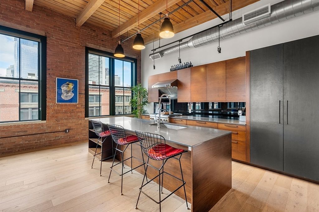This is a modern kitchen with exposed brick walls, wooden beams, and large windows offering city views. The kitchen features sleek wooden cabinetry, a stainless steel range hood, and a large island with bar seating. The overall style is industrial-chic, blending rustic elements with contemporary design.