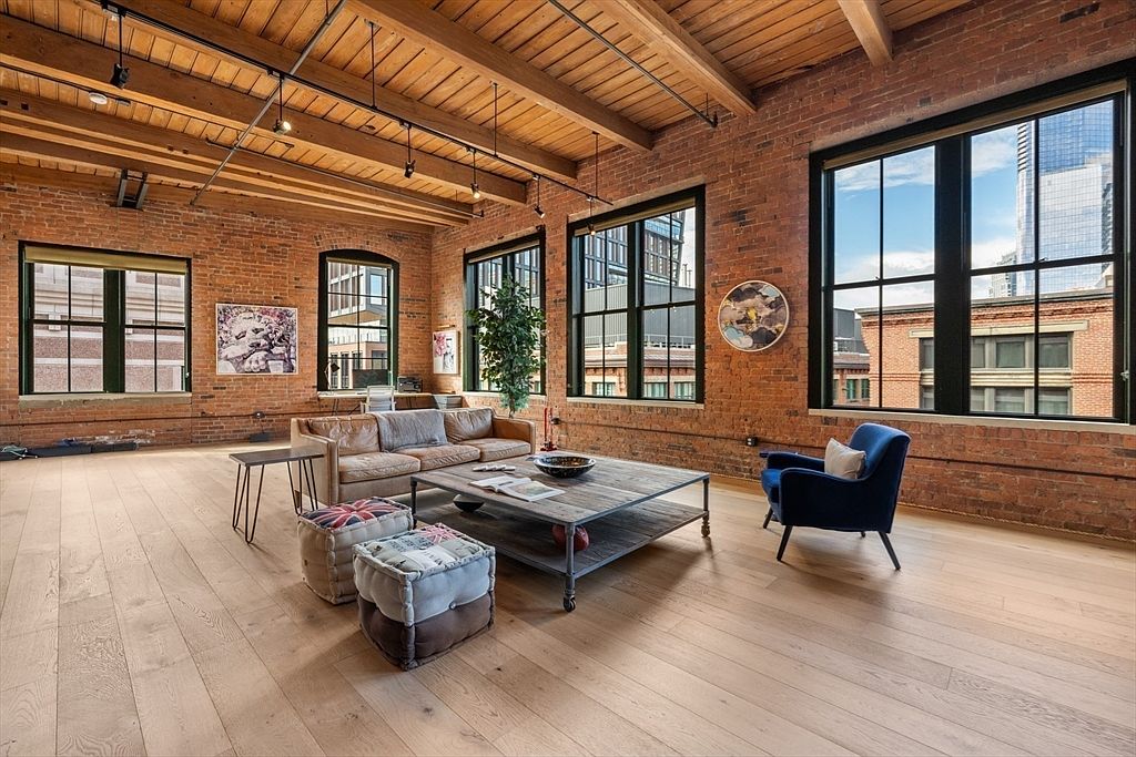 This is a spacious living room with exposed brick walls, wooden beams, and hardwood floors, creating a loft-like atmosphere. Large windows provide ample natural light and city views. The room is furnished with a comfortable sofa, a coffee table, a blue armchair, and decorative ottomans, blending industrial and modern styles.