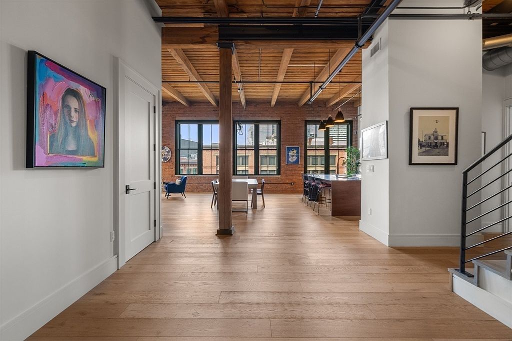 This interior shot showcases a spacious hallway with hardwood flooring leading into an open-concept living area. Exposed brick walls and wooden beams on the ceiling add a rustic, industrial touch. A modern staircase with metal railings is visible on the right, and artwork adorns the walls, creating a stylish and inviting atmosphere.