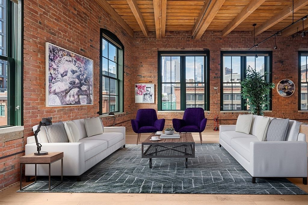 This is an interior shot of a living room featuring exposed brick walls and wooden ceiling beams, creating a loft-like atmosphere. Two white sofas face each other across a patterned rug, with two purple armchairs and a coffee table in between. Large windows provide natural light and city views, enhancing the urban chic aesthetic.