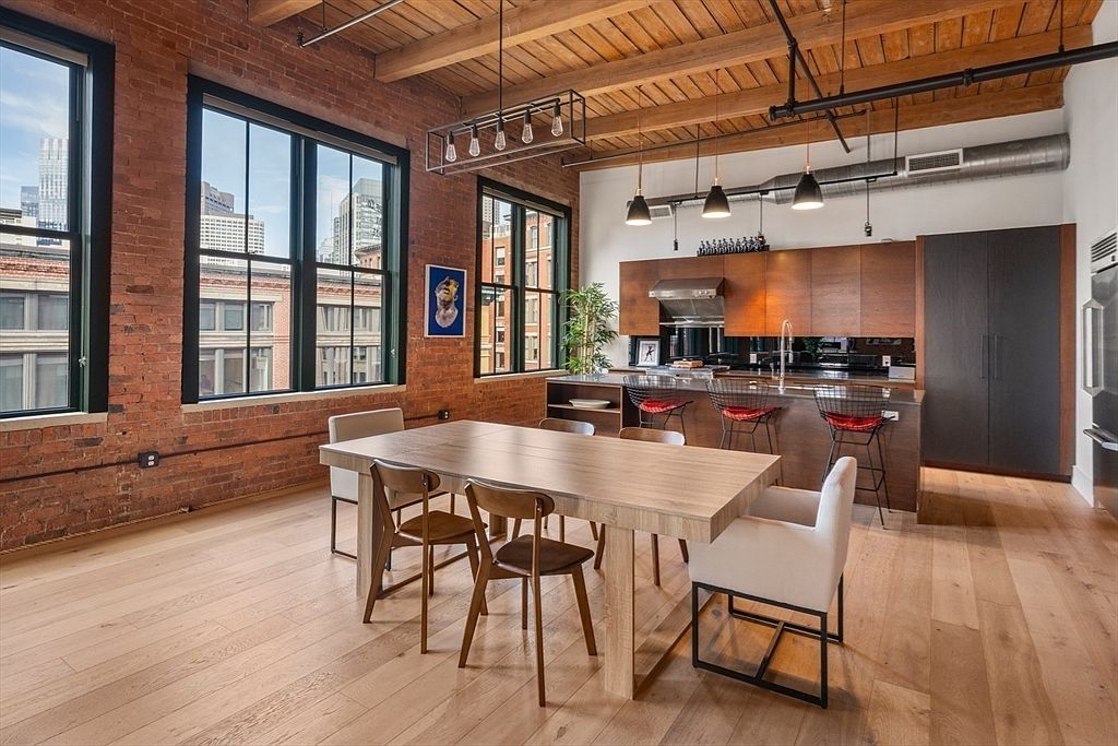 This interior shot showcases a dining area adjacent to a modern kitchen. The dining space features a large wooden table surrounded by a mix of wooden and upholstered chairs, set against a backdrop of exposed brick walls and large windows offering city views. The kitchen boasts sleek, dark wood cabinetry and stainless steel appliances, creating a sophisticated and inviting atmosphere.