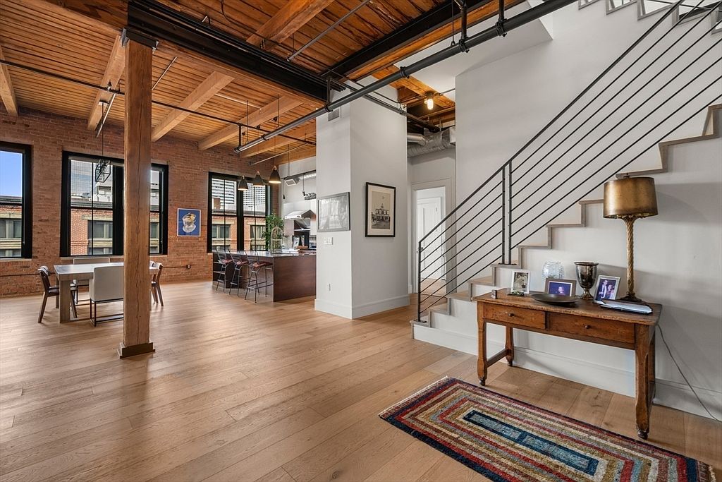 This interior shot showcases a spacious living area with exposed brick walls, wooden beams, and hardwood floors, creating a loft-like atmosphere. The open-concept design integrates a dining area and kitchen, while a staircase with metal railings leads to another level. A rustic wooden console table with a lamp and decorative items adds character to the space.