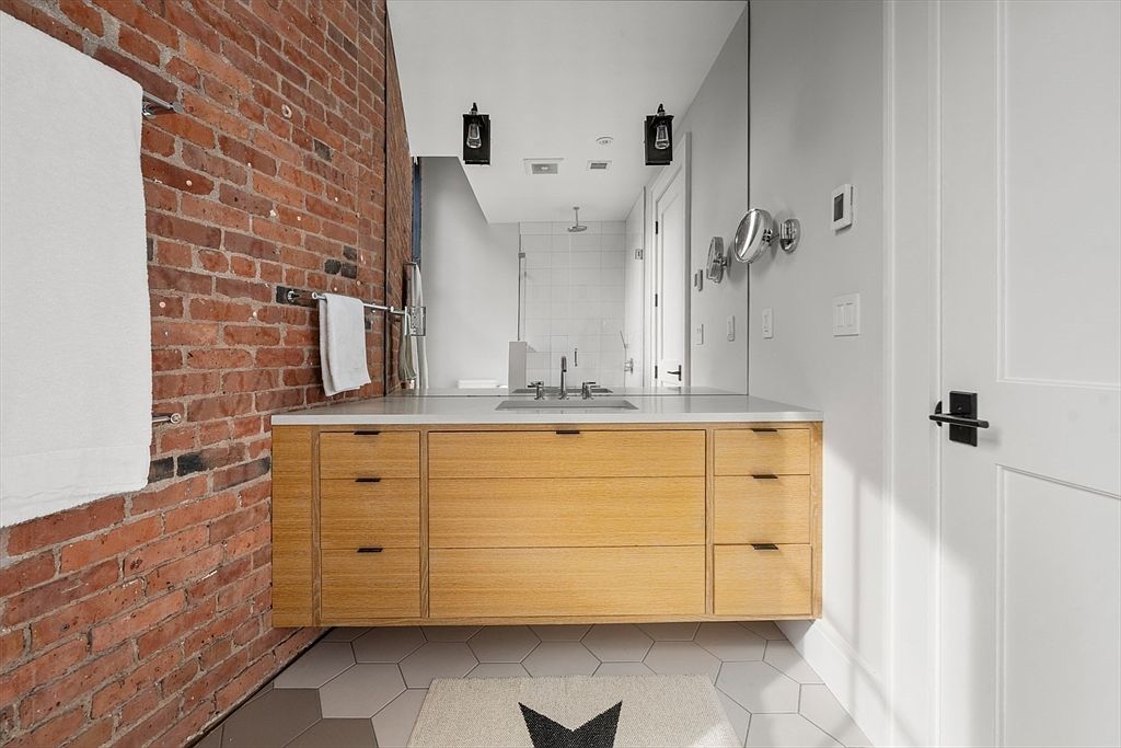 This is a primary bathroom featuring a modern vanity with light wood drawers and a white countertop. A large mirror reflects the rest of the bathroom, including a shower with white tiles. The bathroom has a mix of textures with a brick wall on one side and white hexagonal tiles on the floor, creating a stylish and inviting space.