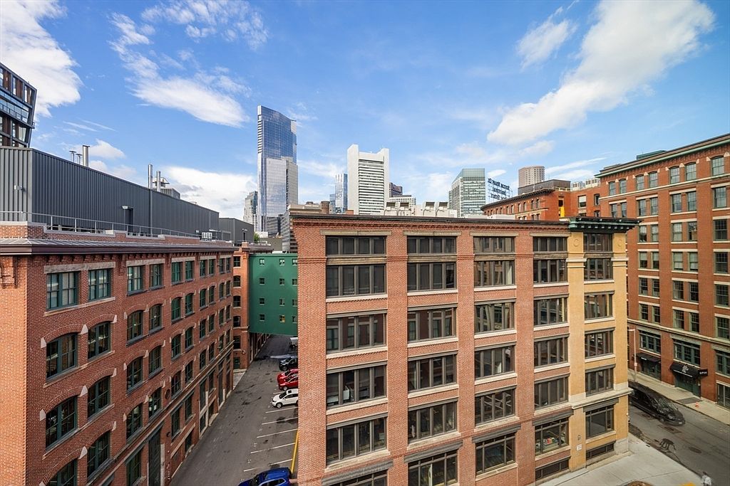 This aerial view showcases a cluster of brick buildings in an urban setting, with a parking lot nestled between them. The cityscape is visible in the background, featuring modern skyscrapers under a partly cloudy sky. The image highlights the building's location and accessibility.