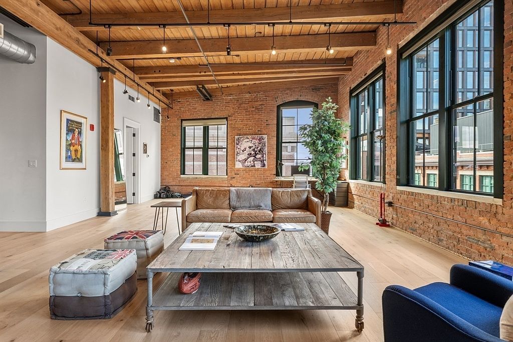 This is an interior shot of a living room featuring exposed brick walls, large windows, and wooden beams on the ceiling, creating a loft-like industrial aesthetic. The room is furnished with a leather sofa, a large wooden coffee table, and decorative ottomans, complemented by a hardwood floor that adds warmth to the space. The perspective is from a medium angle, showcasing the room's spaciousness and architectural details.