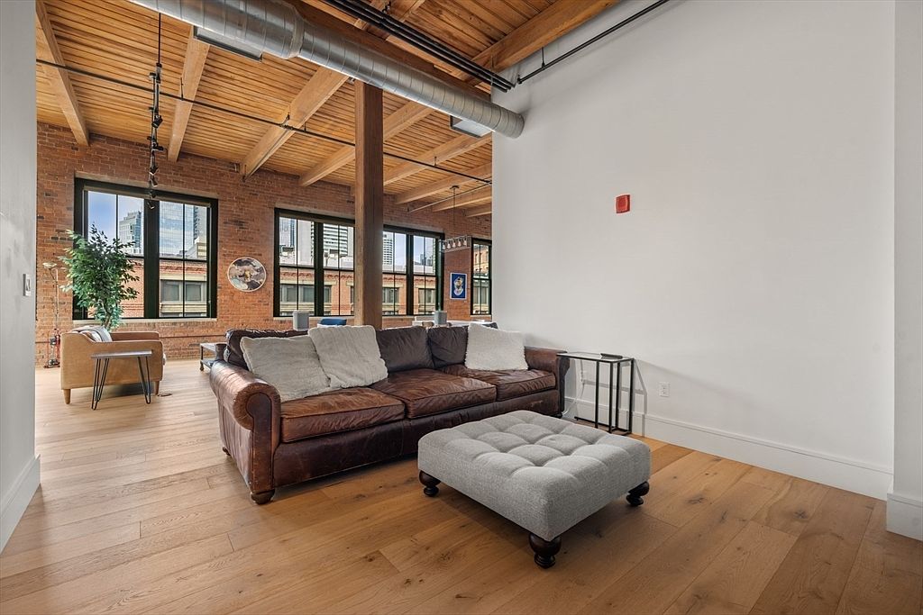 This is an interior shot of a living room featuring a brown leather sofa, a gray ottoman, and hardwood floors. The room has exposed brick walls and wooden beams on the ceiling, giving it a loft-like feel. Large windows provide natural light and views of the city.