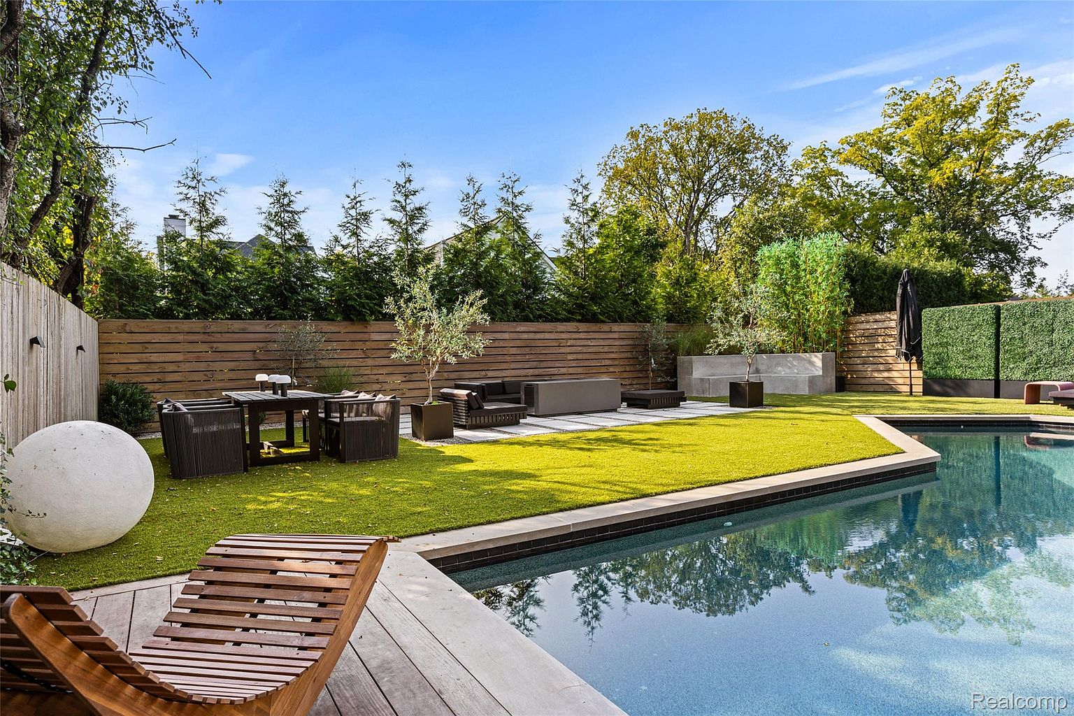 This image showcases a beautifully landscaped backyard featuring a modern design with a pool. The yard includes a well-maintained lawn, a wooden deck with lounge chairs, and a dining area with outdoor furniture. A sleek, contemporary seating area is situated on a paved section, complemented by lush greenery and a wooden fence for privacy, creating an inviting outdoor living space.