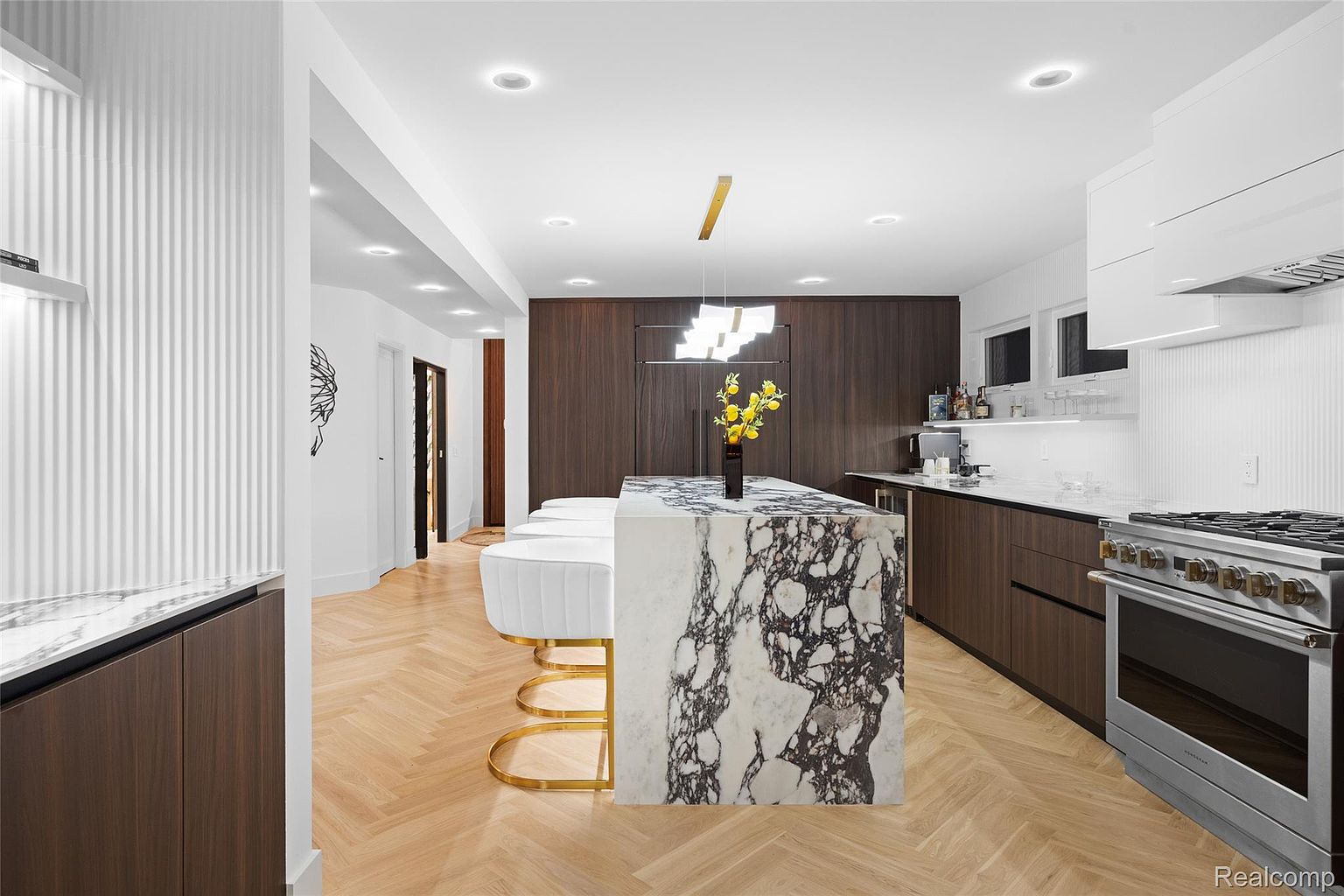 This is a modern kitchen with a large marble island featuring seating and a unique gold base. The kitchen includes dark wood cabinetry, stainless steel appliances, and a herringbone wood floor. The space is well-lit with recessed lighting and a modern chandelier, creating a luxurious and inviting atmosphere.