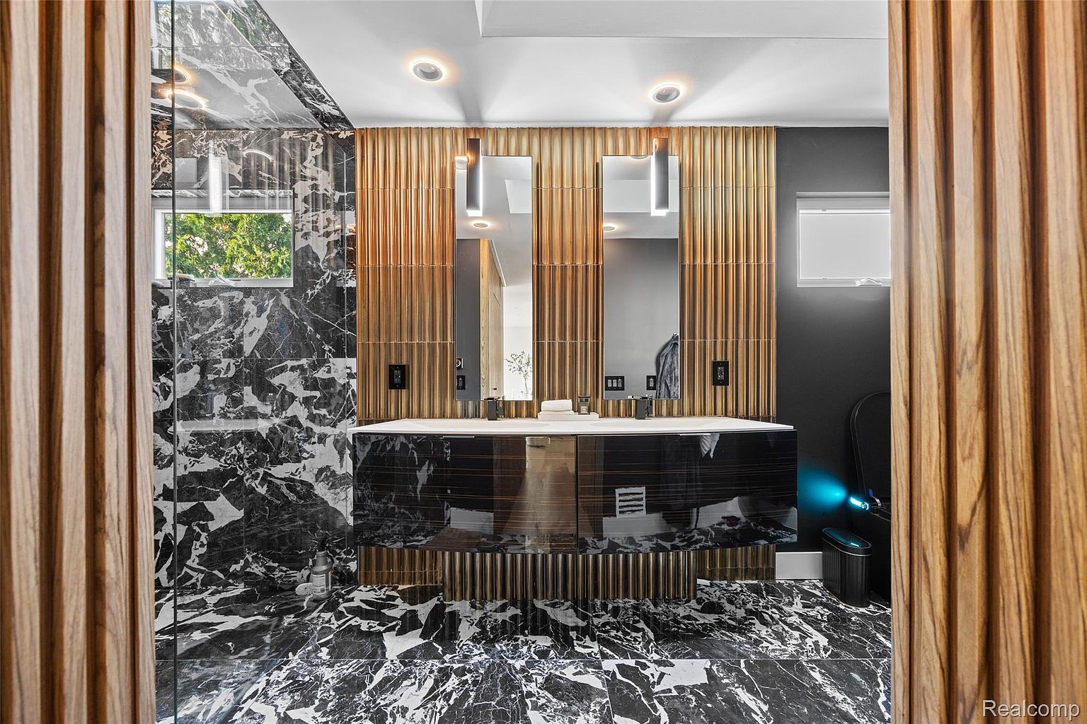 This is a luxurious primary bathroom featuring black and white marble walls and flooring, creating a dramatic and elegant aesthetic. The double vanity has a dark wood finish with gold accents, complemented by two modern mirrors and sleek lighting fixtures. A glass-enclosed shower is visible on the left, and the overall design exudes sophistication and high-end style.