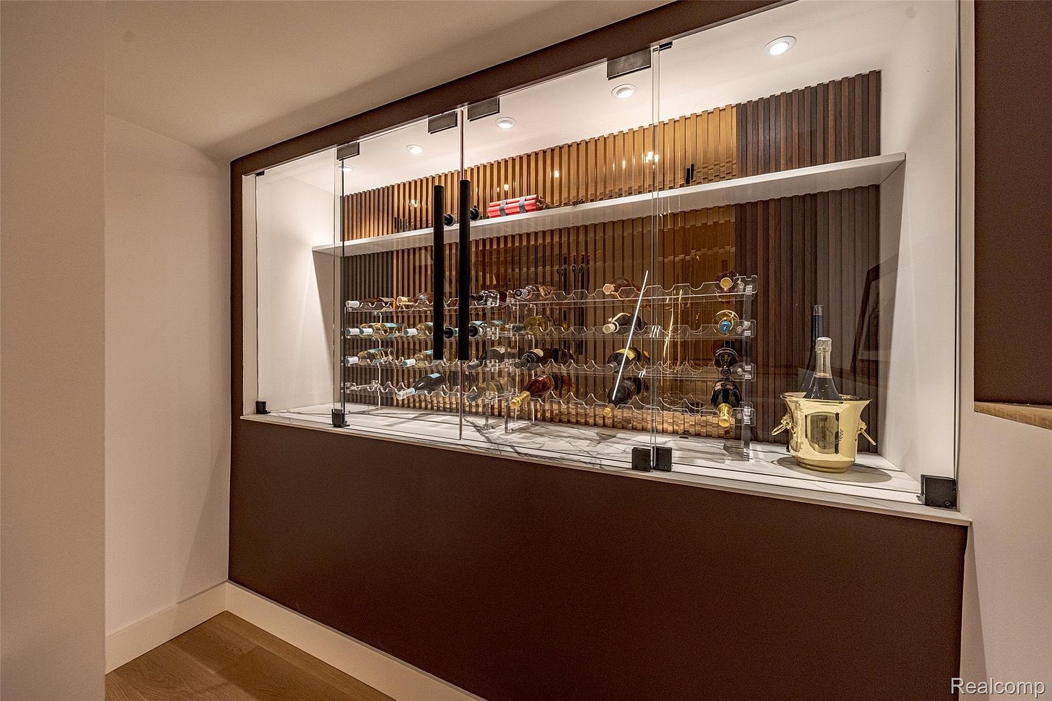 This image showcases a modern wine cellar, featuring a glass-enclosed display with a clear wine rack filled with various wine bottles. The backdrop consists of vertical wooden slats, adding a touch of elegance and warmth. A golden ice bucket with a champagne bottle inside sits on the marble countertop, enhancing the luxurious feel of the space.