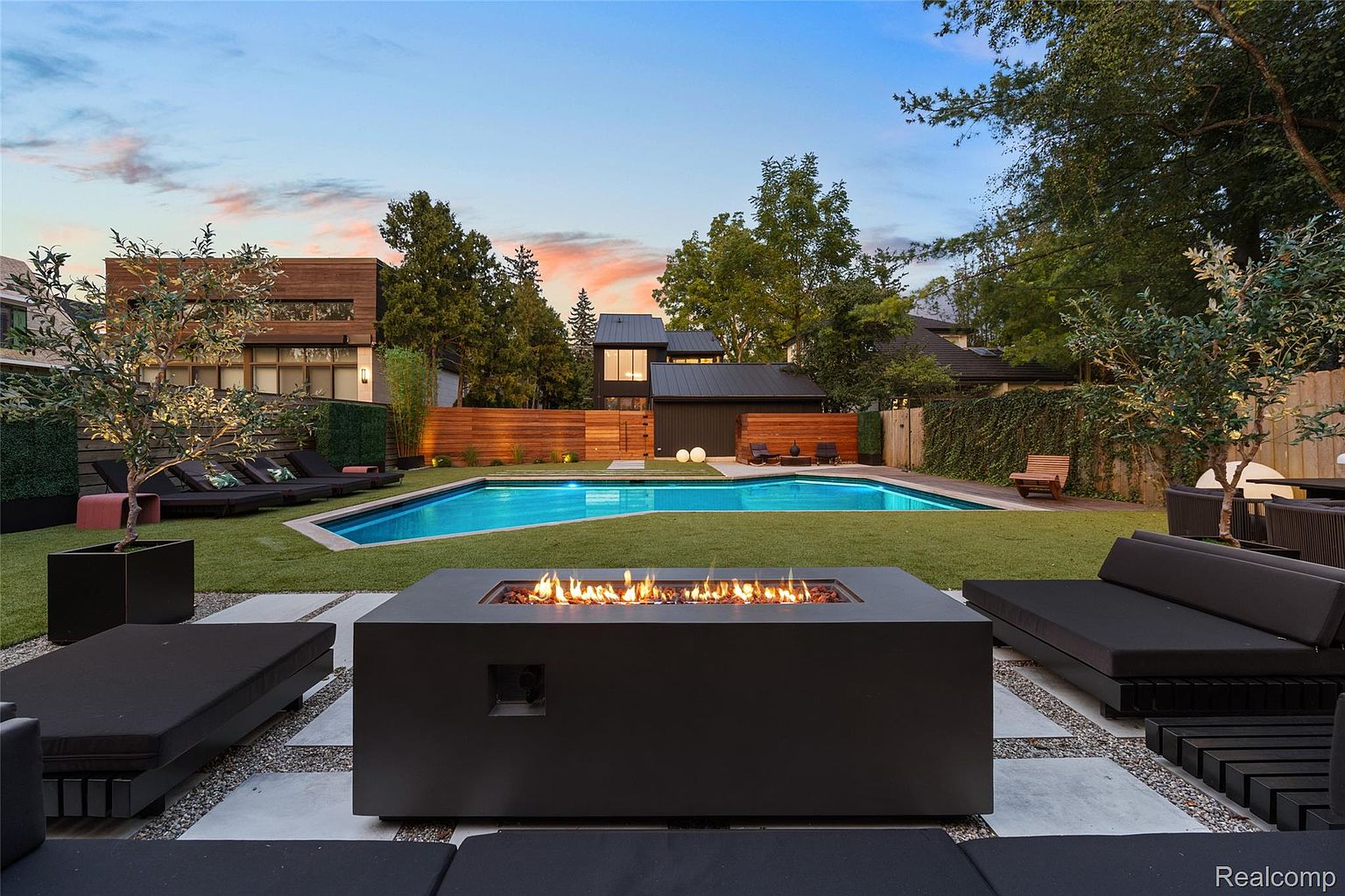 This image showcases a luxurious backyard with a rectangular pool as the central feature. A modern fire pit is surrounded by lounge seating, creating an inviting outdoor living space. The landscape includes manicured grass, trees, and a contemporary home design in the background, suggesting a high-end property.