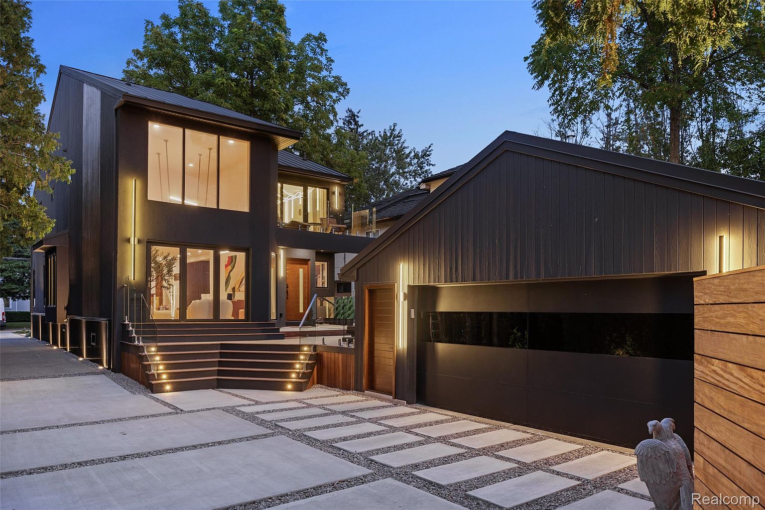 This is a striking front view of a modern home, showcasing its dark exterior, large windows, and well-lit entryway. The property features a multi-level staircase leading to the front door, complemented by a stylish driveway with concrete pavers and gravel accents. The attached garage adds convenience and blends seamlessly with the home's contemporary design.