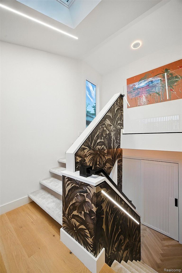 This interior shot showcases a modern staircase with unique design elements. The staircase features carpeted steps, a striking railing adorned with a tropical leaf pattern, and integrated lighting. The walls are painted white, and the flooring is light wood, contributing to a bright and contemporary aesthetic.