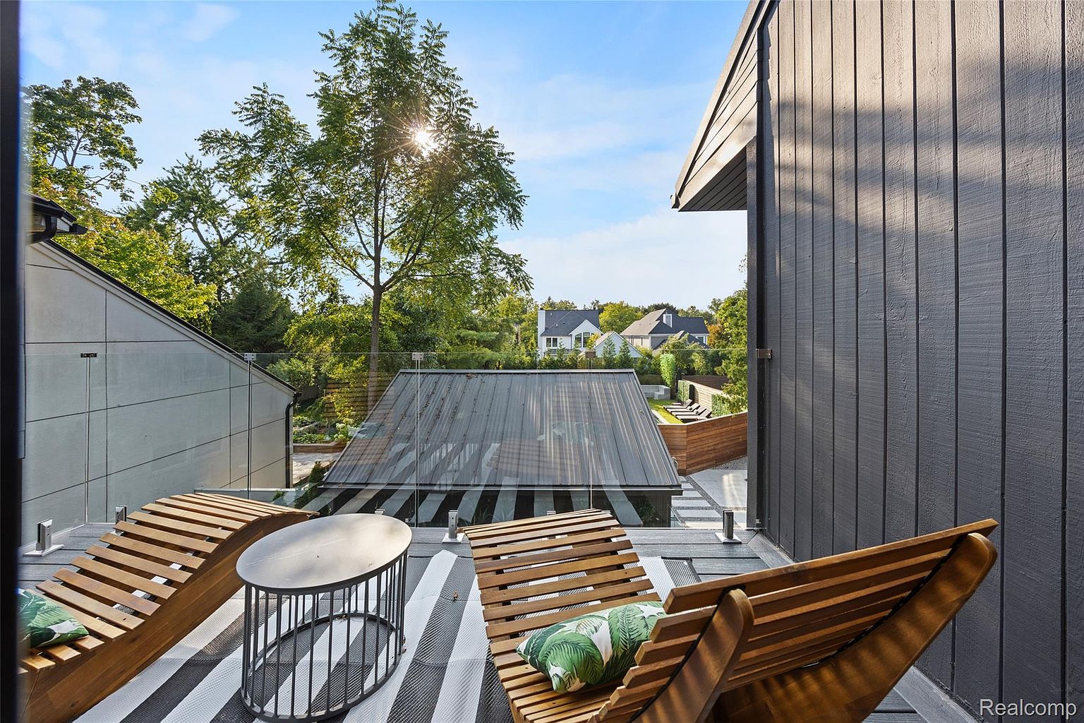 This image showcases a modern balcony or deck area, featuring stylish wooden lounge chairs with cushions and a small round table on a black and white striped rug. A glass railing provides an unobstructed view of the surrounding greenery and neighboring houses, while the dark gray siding of the building adds a contemporary touch. The space is designed for relaxation and outdoor enjoyment.