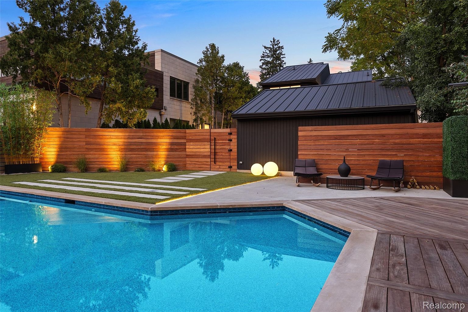This image showcases a luxurious backyard pool area, featuring a modern pool with a tiled interior and a concrete surround. A wooden deck extends from the pool area, furnished with stylish outdoor seating. The space is enclosed by a contemporary wooden fence, complemented by ambient lighting and lush greenery, creating a private and sophisticated outdoor retreat.