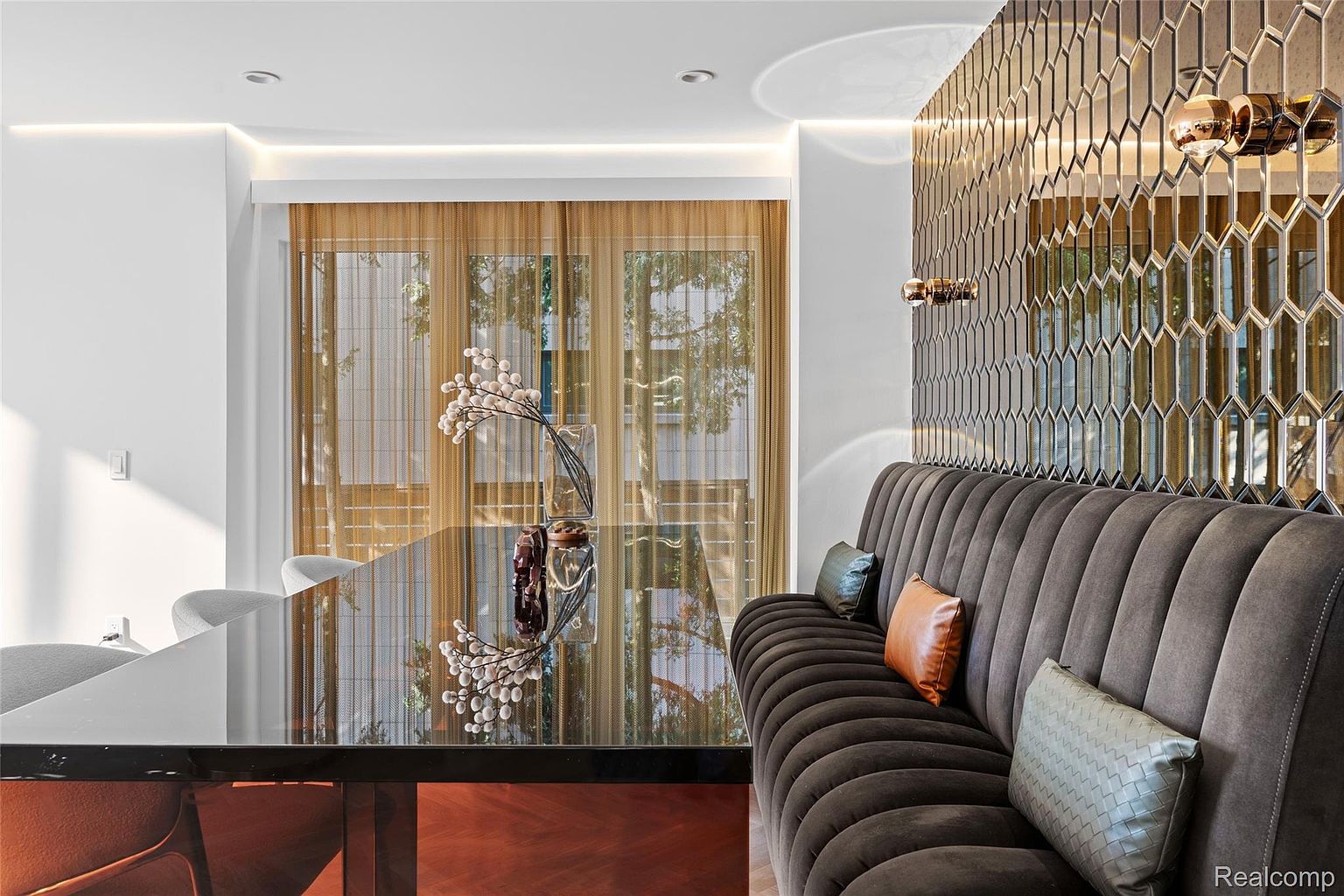 This image showcases a stylish dining room featuring a dark, reflective dining table adorned with a floral arrangement. A plush, tufted banquette lines the wall, complemented by decorative pillows and a mirrored geometric wall design with sconces. The room exudes a modern and sophisticated ambiance.