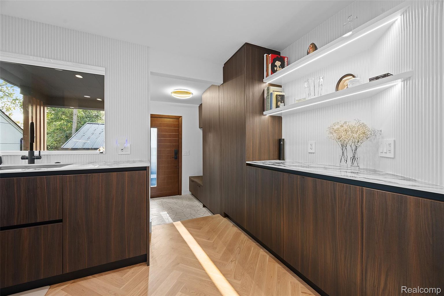 This interior shot showcases a modern kitchen with dark wood cabinetry and a light-colored countertop. The space features built-in shelving with integrated lighting, adding a touch of elegance. The herringbone wood flooring and the view to the outside through a window contribute to a bright and inviting atmosphere.