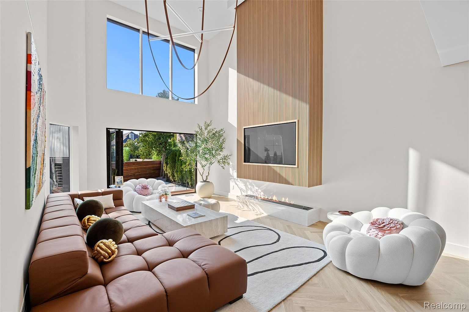This is a bright and modern living room featuring a large brown sectional sofa, two unique white pouf chairs, and a sleek fireplace. The room has high ceilings, a large window, and a statement wood-paneled wall with a mounted television. The overall aesthetic is luxurious and contemporary, creating a welcoming and stylish space.