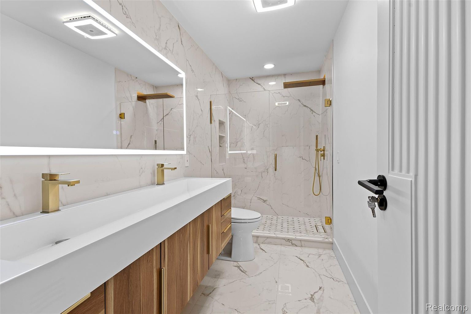 This is a modern primary bathroom featuring marble-style tile on the walls and floors, a double vanity with a white countertop and wood-look cabinets, and gold-tone fixtures. A glass-enclosed shower with gold hardware is visible, along with a toilet. The overall impression is luxurious and contemporary, with a focus on clean lines and high-end finishes.