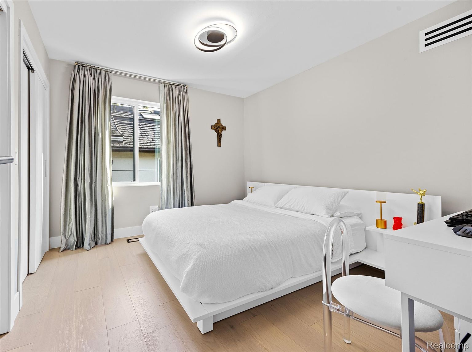This is a bright and airy bedroom featuring a minimalist design with a white platform bed, white bedding, and light wood flooring. The room has a window with silver curtains, a modern ceiling light, and a white desk with a clear chair. A wooden cross hangs on the wall, adding a touch of traditional decor.