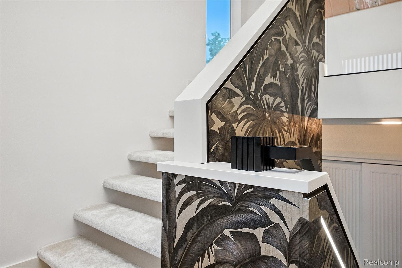 This interior shot showcases a modern staircase with carpeted steps and a unique wallpaper featuring a tropical leaf pattern on the side walls. The staircase is painted white, providing a clean contrast to the patterned wallpaper. A black decorative object sits atop the railing, adding a touch of contemporary style.