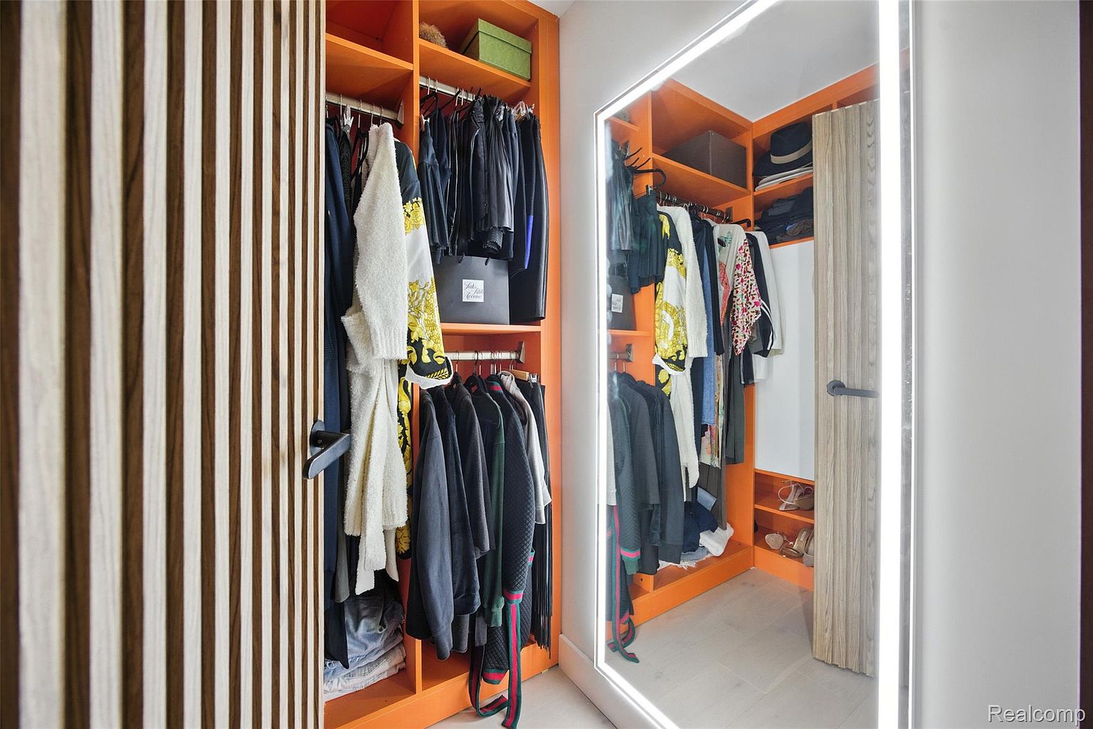 This image showcases a well-organized walk-in closet with orange shelving and hanging rods filled with clothing. A full-length mirror with integrated lighting adds a modern touch and enhances the space. The closet features a mix of open shelving and enclosed storage, providing ample space for clothes and accessories.