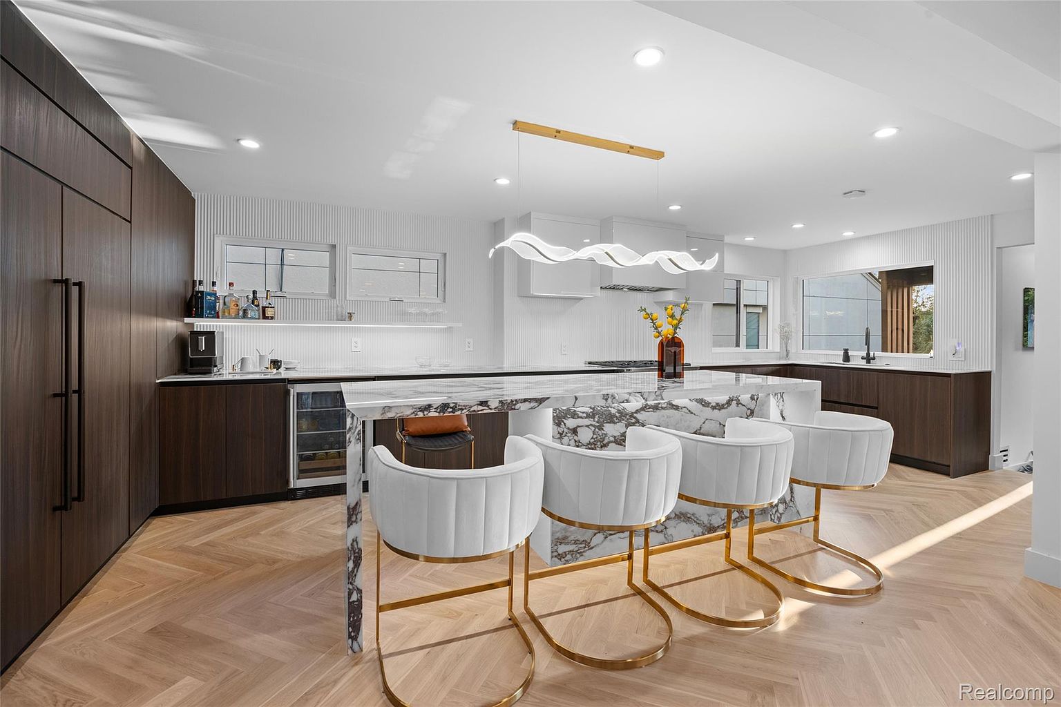 This is a bright, modern kitchen featuring a large island with marble countertops and waterfall edges, complemented by stylish white bar stools with gold accents. The kitchen includes dark wood cabinetry, stainless steel appliances, and unique lighting fixtures, creating a luxurious and inviting space. The herringbone wood flooring adds a touch of elegance.