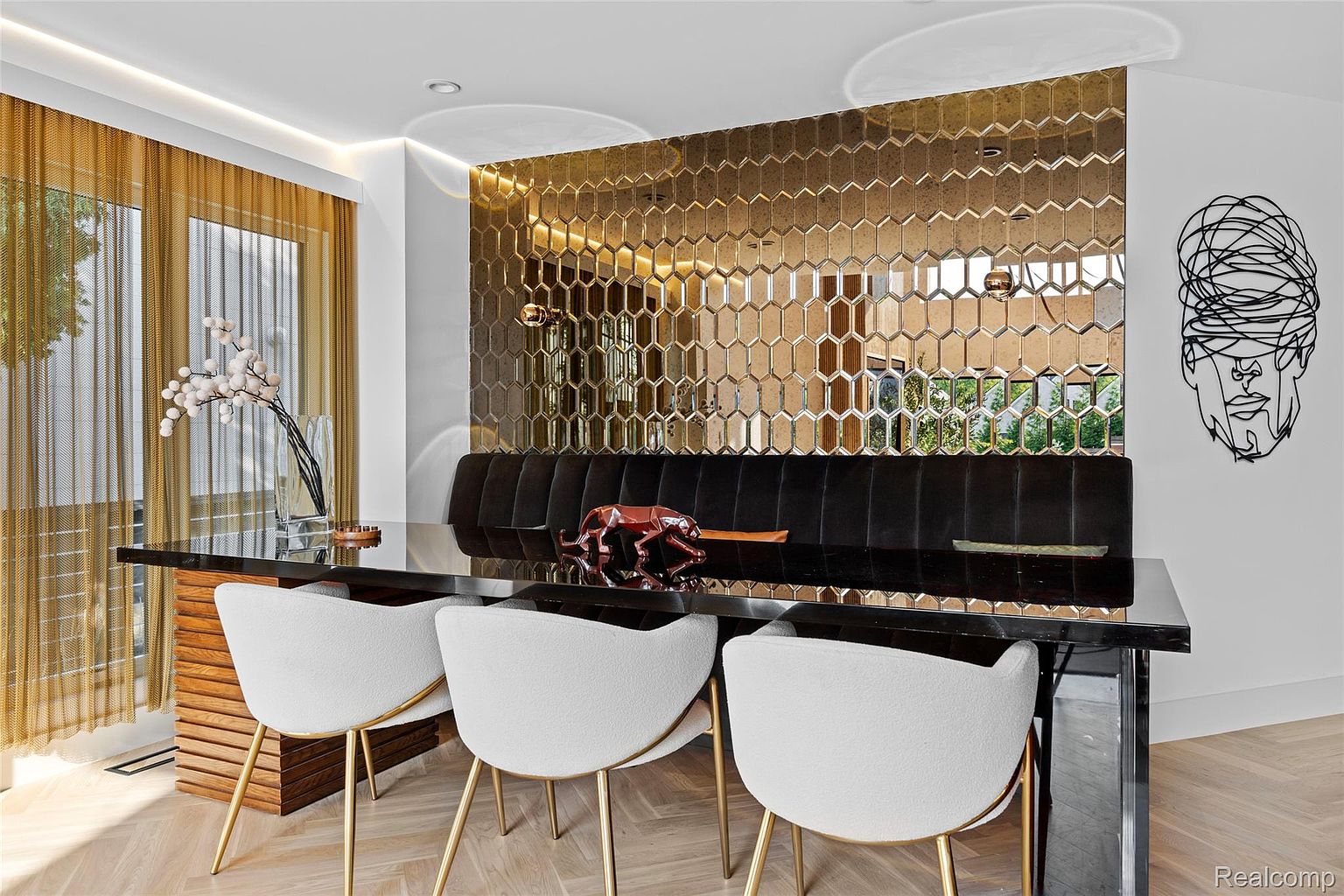 This dining room features a modern aesthetic with a black glass-topped table, complemented by white chairs with gold legs. A striking gold honeycomb-patterned wall adds texture and visual interest, while a black velvet banquette provides additional seating. The room is well-lit and stylish, creating an inviting space for dining and entertaining.