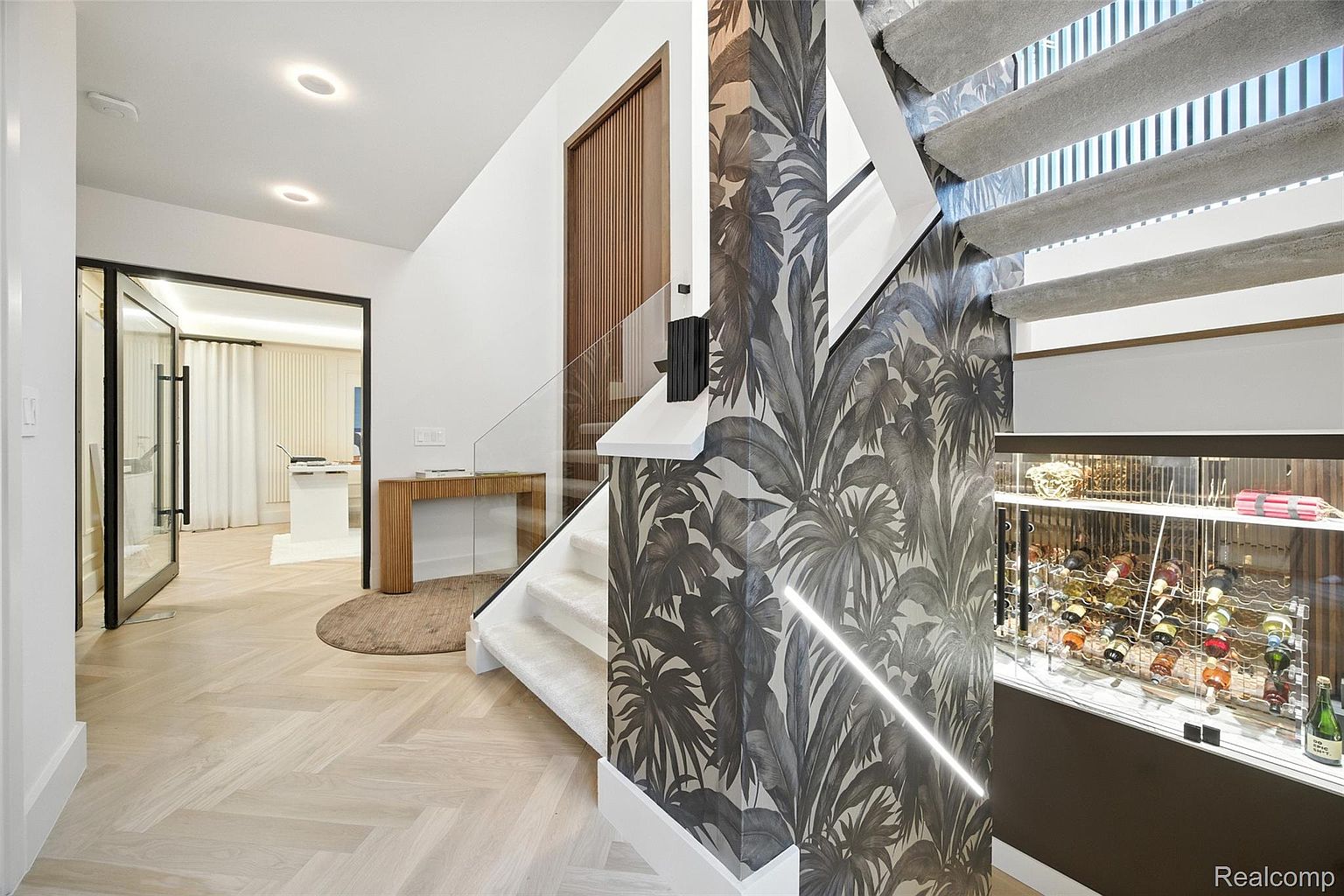 This interior shot showcases a modern hallway with a striking staircase featuring tropical-themed wallpaper and glass railings. The herringbone wood flooring adds a touch of elegance, while the adjacent room is visible through a glass door. A built-in wine cellar is visible under the stairs.