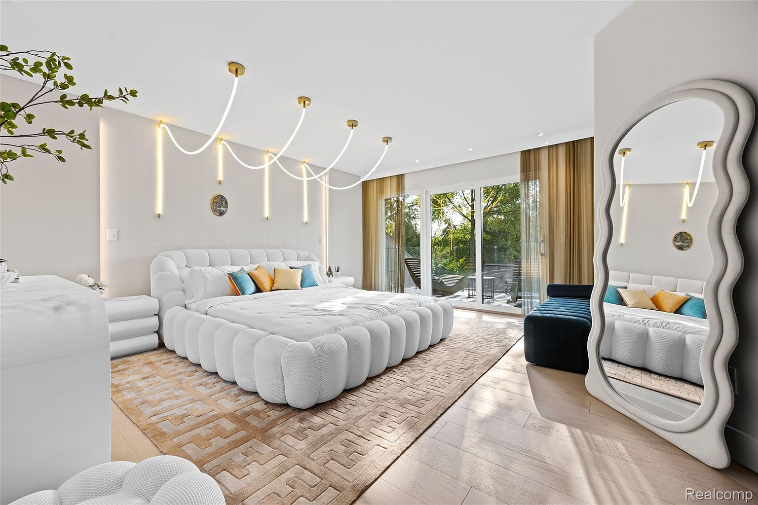 This is a bright and airy primary bedroom featuring a unique, oversized white upholstered bed with matching side tables. The room is decorated with modern lighting fixtures and a large, organically shaped mirror. A sliding glass door leads to an outdoor balcony, enhancing the sense of space and luxury.