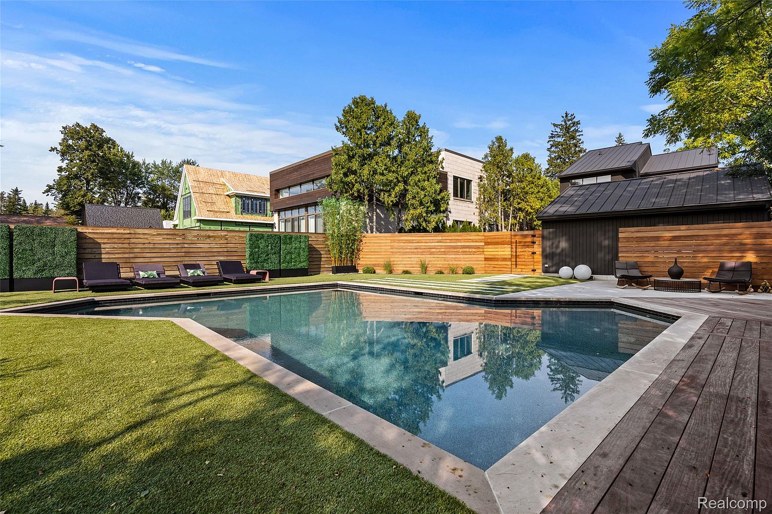 This image showcases a luxurious backyard pool area. The pool features a modern design with clean lines and is surrounded by a well-maintained lawn, a wooden deck, and a stylish fence for privacy. Contemporary lounge chairs and decorative elements enhance the upscale ambiance, making it an ideal outdoor space for relaxation and entertainment.