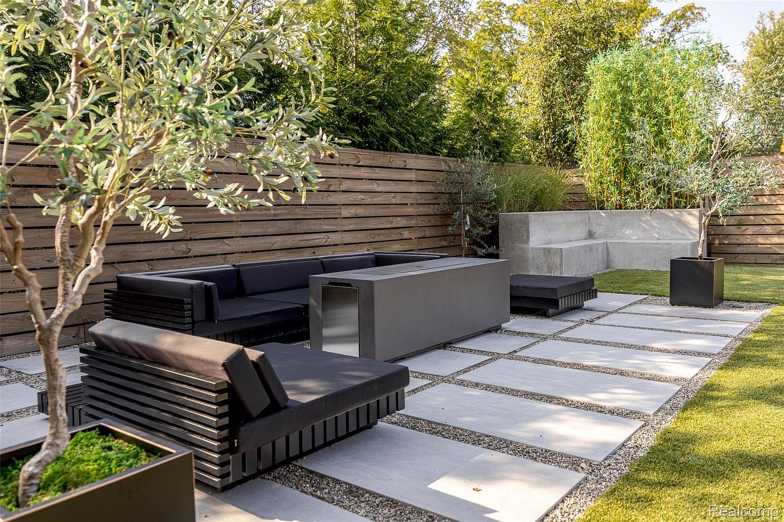 This is a beautifully designed backyard featuring a modern outdoor living space. The area includes a sleek, dark-colored sectional sofa and lounge chair arranged around a contemporary fire pit. The patio is constructed with large, light-colored pavers separated by gravel, and the space is surrounded by lush greenery and a wooden fence, creating a private and inviting atmosphere.