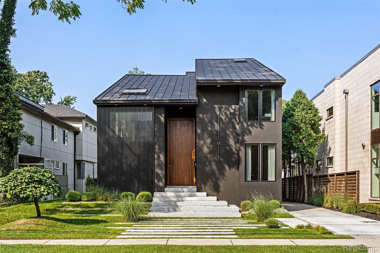 This is a striking front view of a modern home with a dark exterior, featuring a unique architectural design. The house has a metal roof, a wooden front door, and a minimalist facade with vertical slat accents. The landscaping includes a well-manicured lawn, stepping stone pathway, and carefully placed shrubbery, enhancing the property's curb appeal.