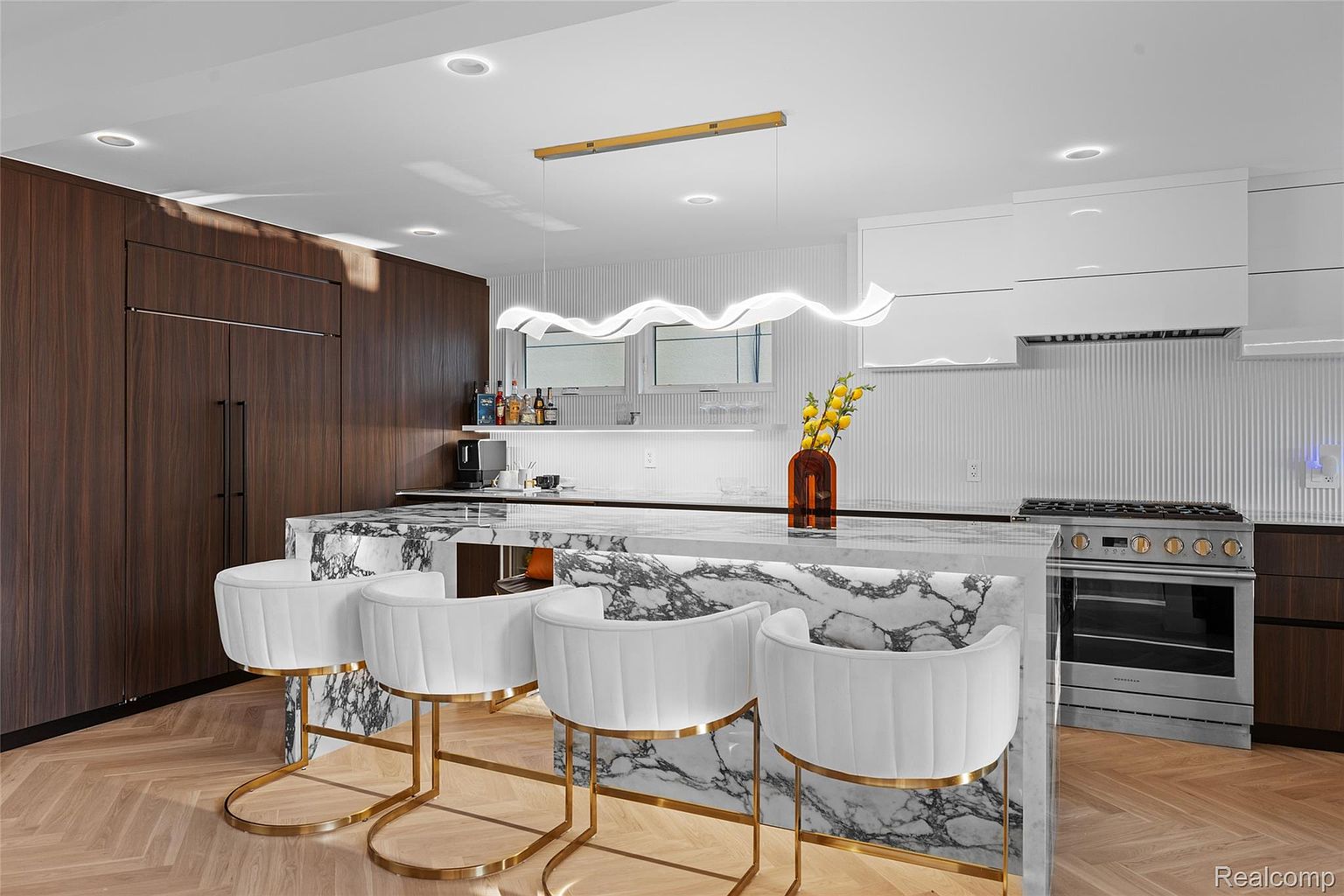 This is a modern kitchen featuring a marble island with seating, dark wood cabinetry, and stainless steel appliances. The island is topped with a marble countertop and accented by gold-legged white chairs. A unique, wavy light fixture hangs above the island, adding a touch of contemporary elegance to the space.