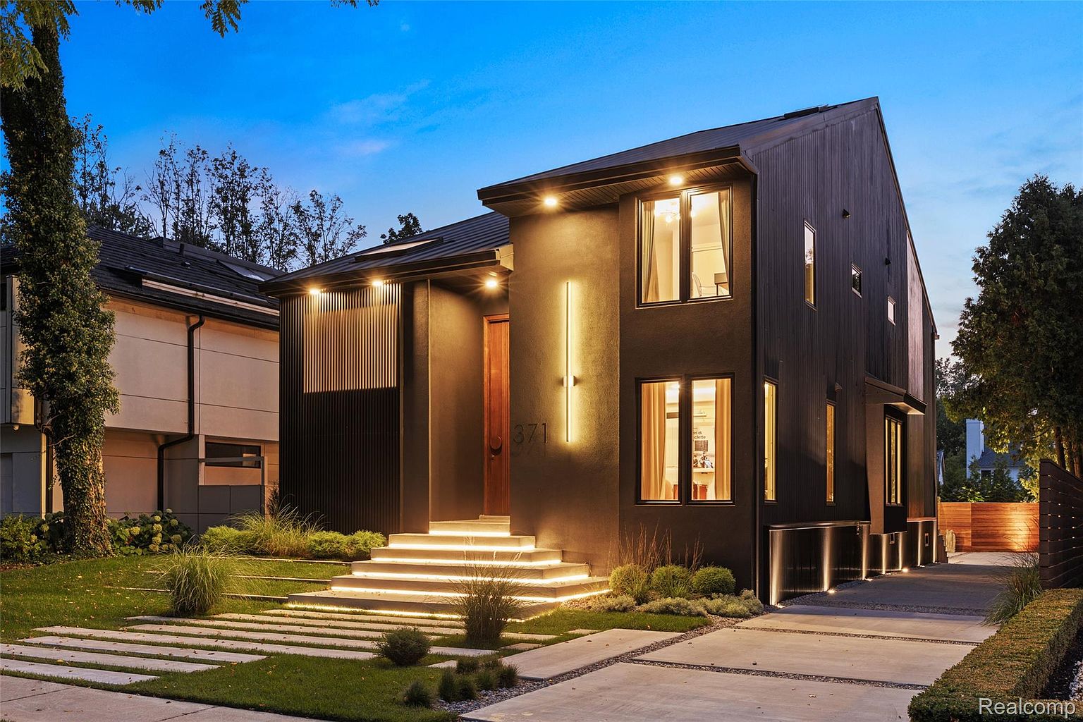 This is a striking front view of a modern home at dusk, showcasing its unique architectural design. The exterior features a dark facade, illuminated entryway with glowing steps, and contemporary landscaping. The clean lines and minimalist aesthetic create a sophisticated and inviting impression.