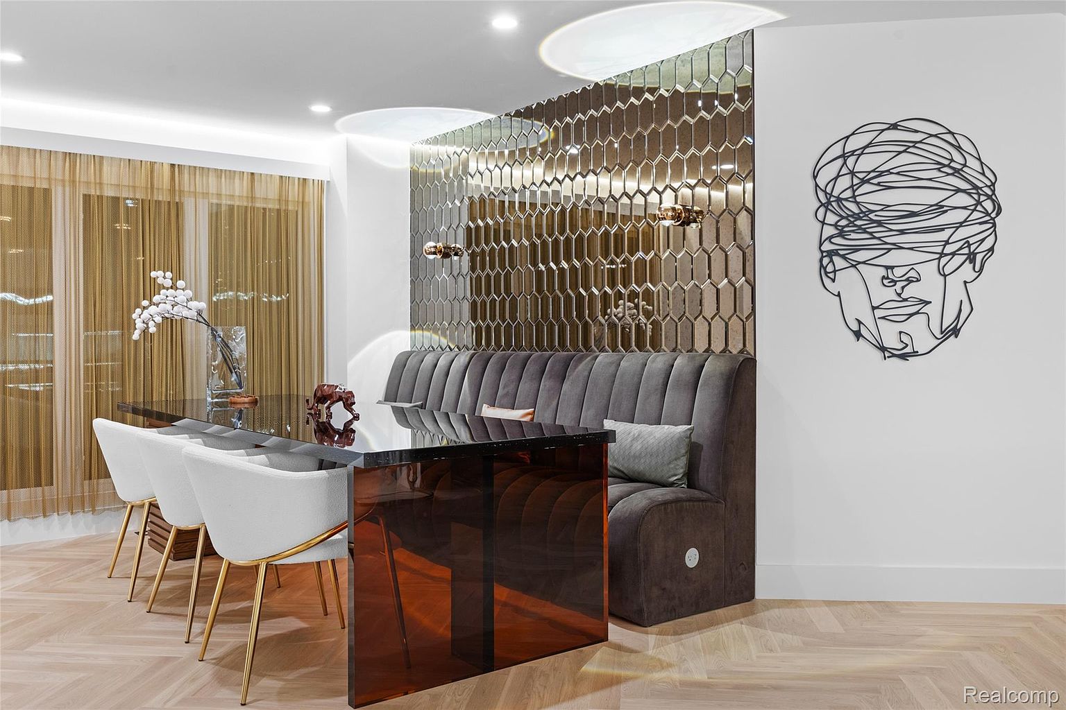 This is an interior shot of a dining room featuring a modern aesthetic. The room includes a dark glass dining table surrounded by white chairs with gold legs and a built-in gray banquette. A mirrored wall with geometric patterns adds visual interest, complemented by a unique piece of art on the adjacent wall, all set against a light wood floor.