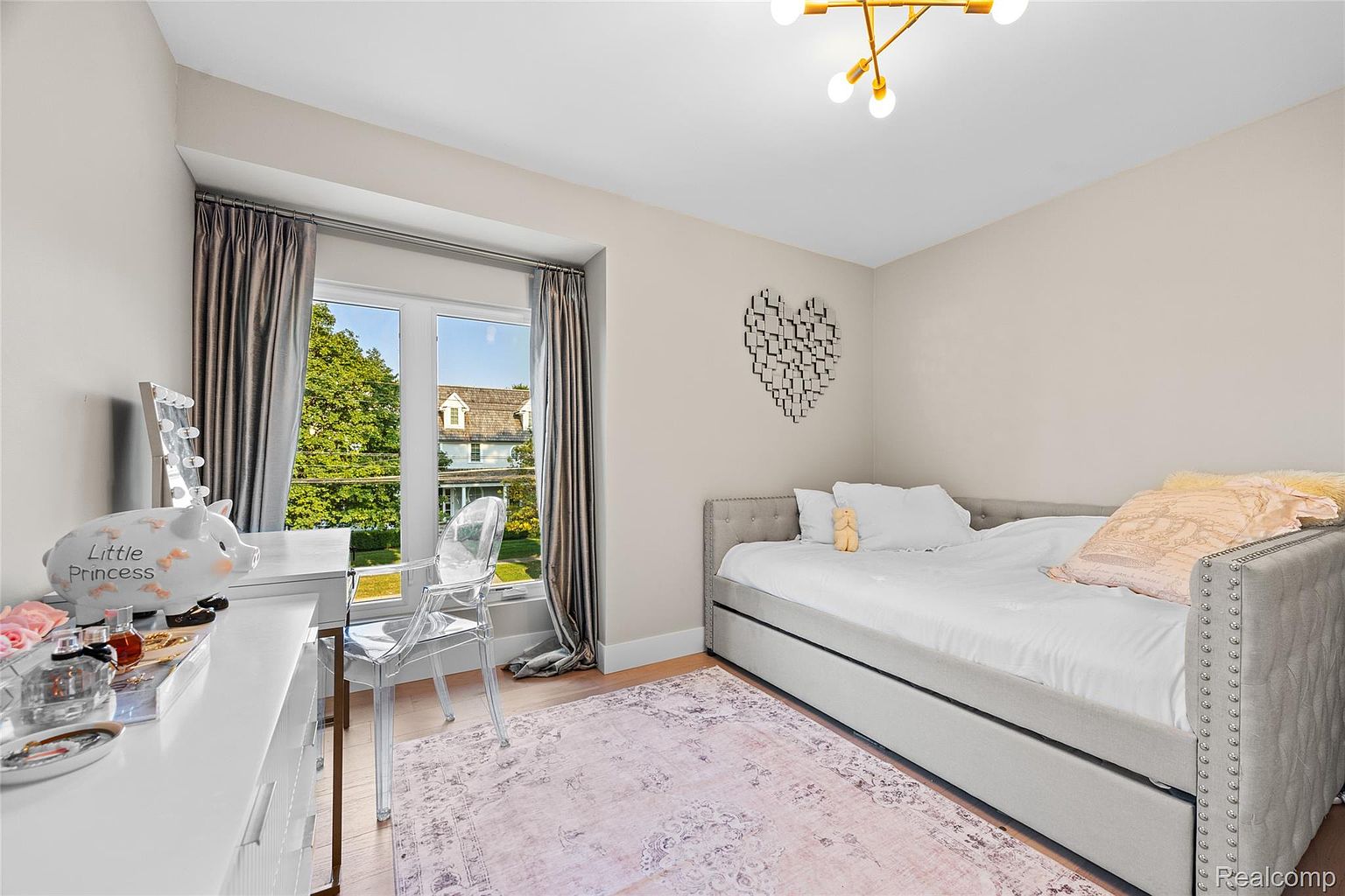 This is a charming bedroom featuring a daybed with a gray upholstered frame and nailhead trim, adorned with white linens and decorative pillows. A clear acrylic chair sits beside a white desk, positioned in front of a window with gray curtains, offering a view of the outdoors. The room is decorated with a heart-shaped wall art and a pink patterned rug, creating a cozy and inviting atmosphere.