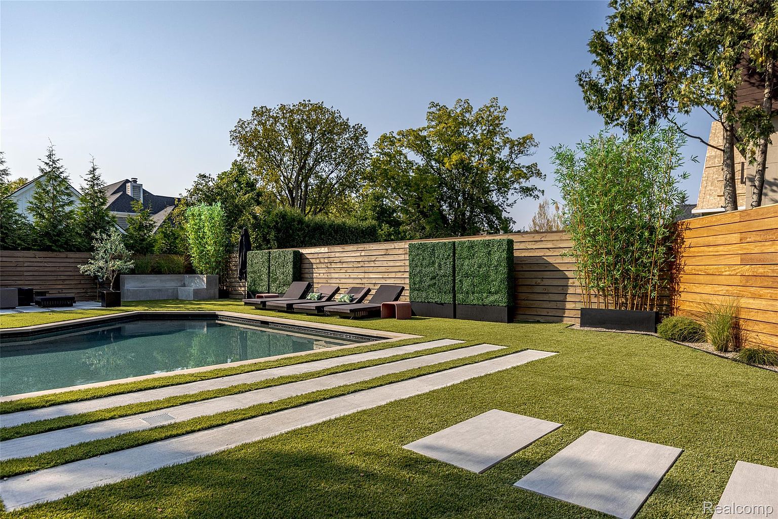 This image showcases a modern backyard with a rectangular pool, surrounded by artificial turf and concrete pavers. A wooden fence provides privacy, complemented by strategically placed greenery and lounge chairs. The scene exudes a luxurious and serene atmosphere, perfect for relaxation and entertainment.