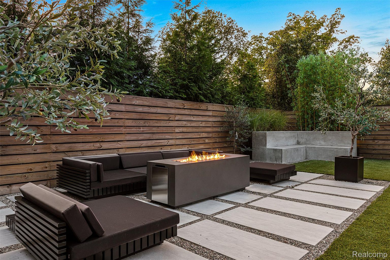 This image showcases a modern outdoor patio area, featuring a sleek fire pit as the focal point, surrounded by contemporary lounge seating. The patio is paved with large square stones interspersed with gravel, creating a clean and stylish look. A wooden fence provides privacy, while lush greenery and trees in the background enhance the serene atmosphere, making it an ideal space for relaxation and entertaining.