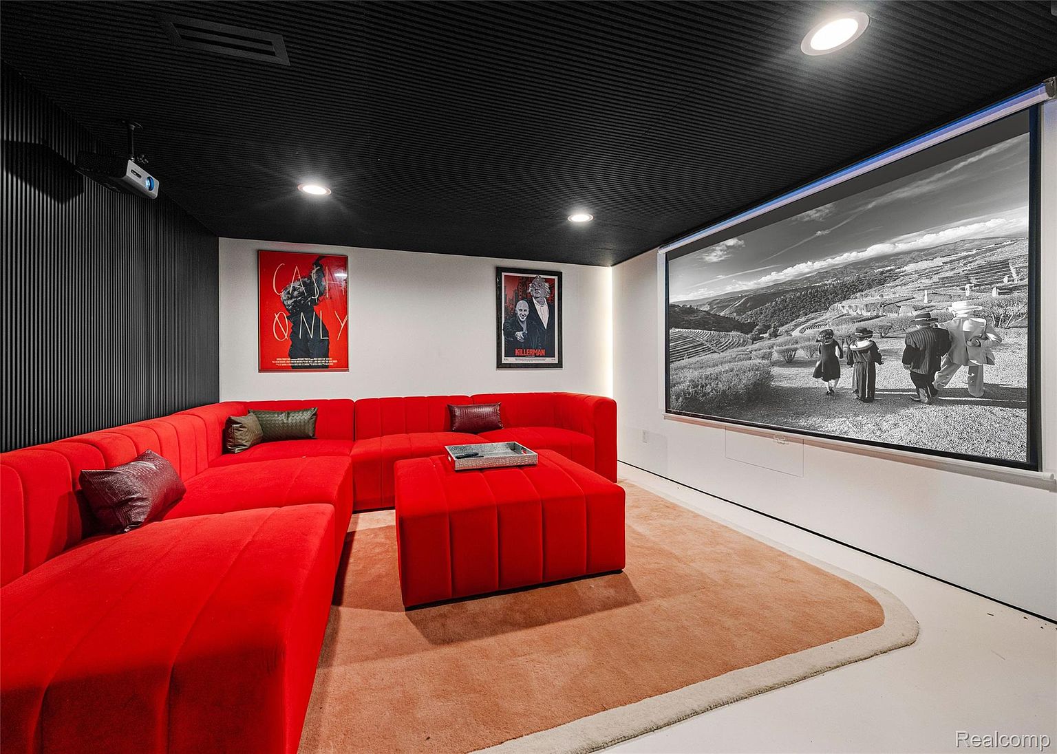 This is a modern home theater room featuring a large red sectional sofa and ottoman, creating a comfortable and inviting space. A projector screen displays a black and white film, while movie posters adorn the walls. The room is illuminated by recessed lighting and a projector, enhancing the cinematic experience.