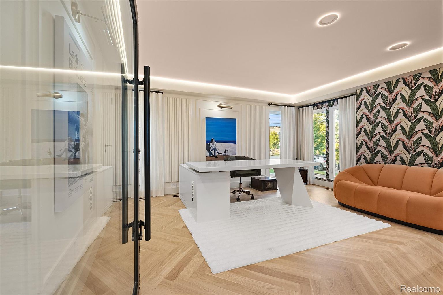 This is an interior shot of a modern office space featuring a large white desk, a comfortable office chair, and a stylish orange sofa. The room is well-lit with recessed lighting and natural light from the windows, which are framed by elegant curtains. The flooring is a light wood with a herringbone pattern, and a patterned wallpaper adds a touch of sophistication to the space.