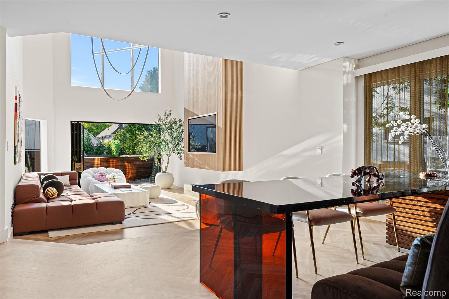 This is a bright and airy living room featuring modern design elements. The room includes a brown leather sofa, a unique white armchair, and a sleek coffee table, all set on a light wood floor. A large window and an open doorway provide ample natural light and a view to the outside, while a wooden feature wall with a mounted TV adds a touch of sophistication.