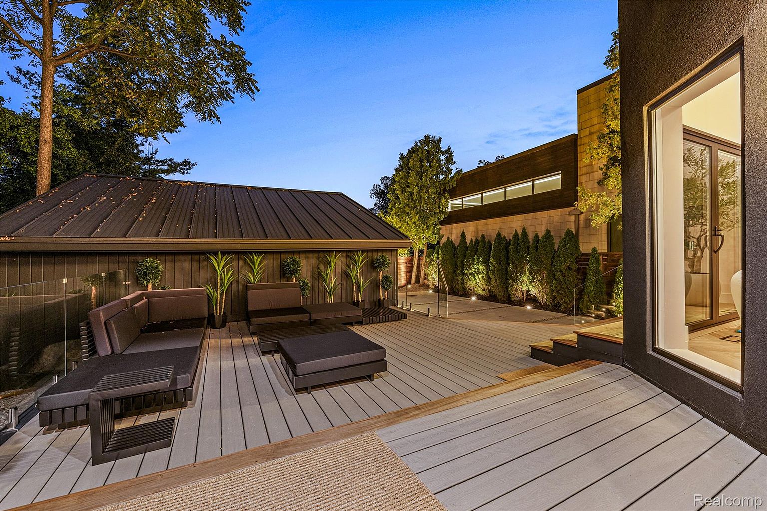 This image showcases a modern outdoor patio and deck area. The space features sleek, dark-toned outdoor furniture, including a sectional sofa and ottomans, arranged on a gray composite deck. A covered structure with a dark metal roof provides shade, while manicured greenery adds a touch of nature. The overall impression is one of sophisticated outdoor living and relaxation.