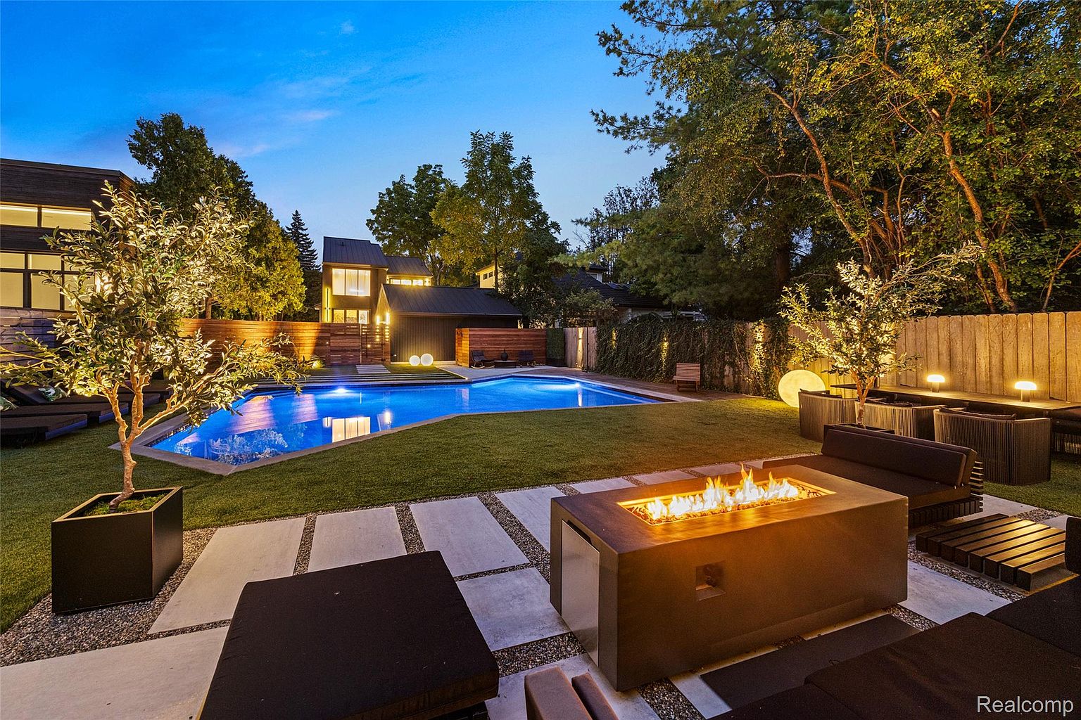This image showcases a luxurious backyard featuring a modern swimming pool with a cover, surrounded by lush green grass and stylish landscaping. A contemporary fire pit with seating area adds a cozy touch, while the well-lit trees and architectural lighting enhance the ambiance. The scene exudes a sense of relaxation and upscale outdoor living.
