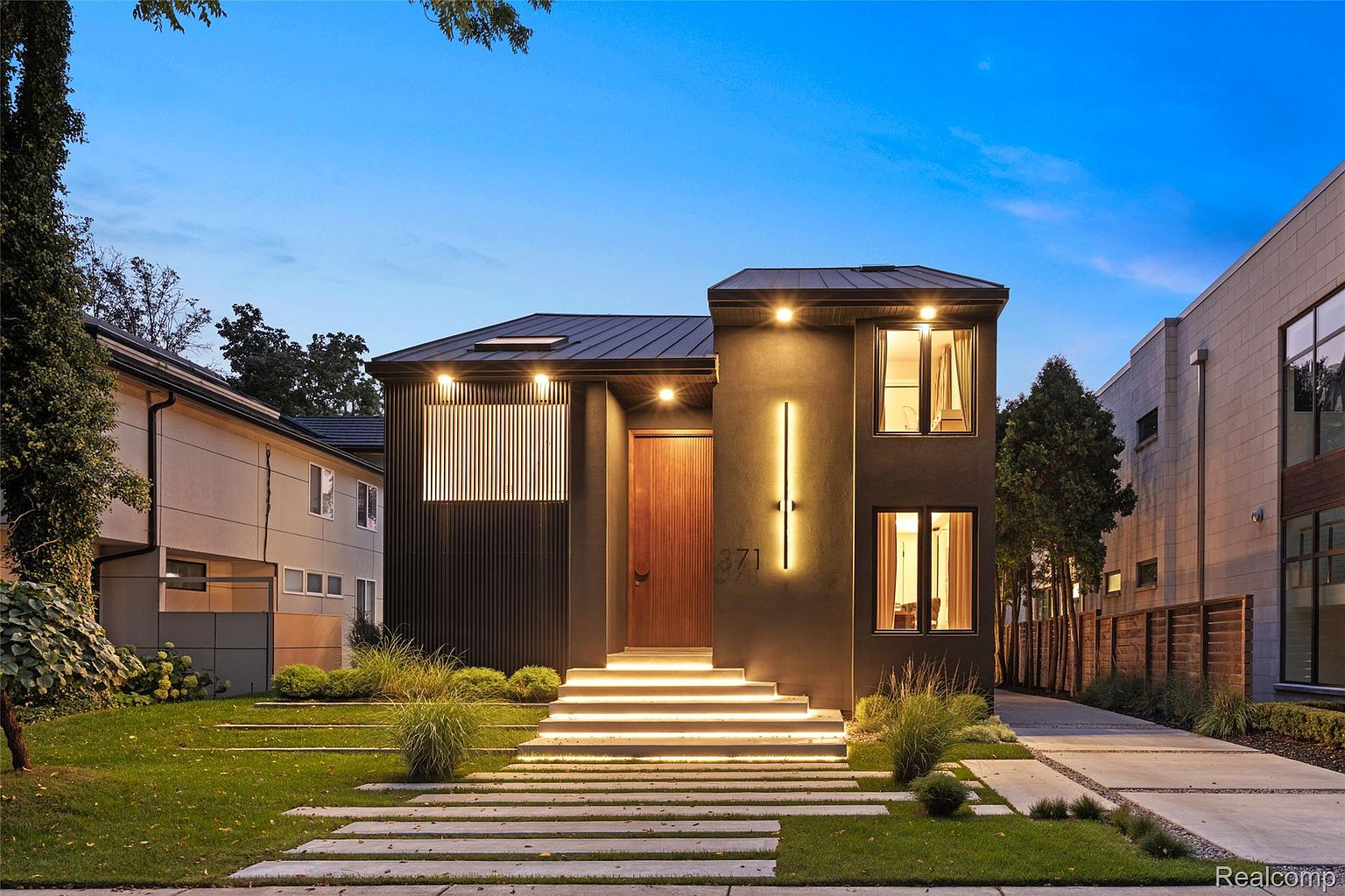 This is a front exterior view of a modern home at dusk. The house features a dark facade, a wooden front door, and illuminated steps leading to the entrance. The landscaping includes a well-manicured lawn and strategically placed shrubbery, enhancing the property's curb appeal.